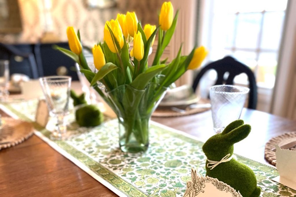 a spring themed tablescape with fresh yellow tulips and a green and white runner on the middle of the table, as well as faux moss bunnies for decoration